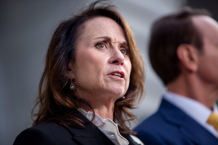 A woman with shoulder-length brown hair wearing pearl earrings, a black blazer and a white ruffled shirt looks off camera. She is standing in front of a man wearing a blue suit and yellow tie. News microphones appear in the lower right corner.
