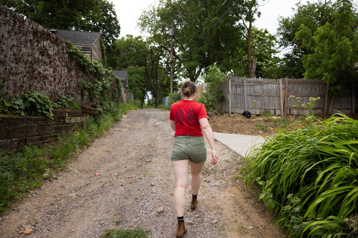 A woman wearing a red T-shirt, green shorts and brown boots walks down a dirt path. Bushes and grass line the sides of the path, and trees grow in the distance.