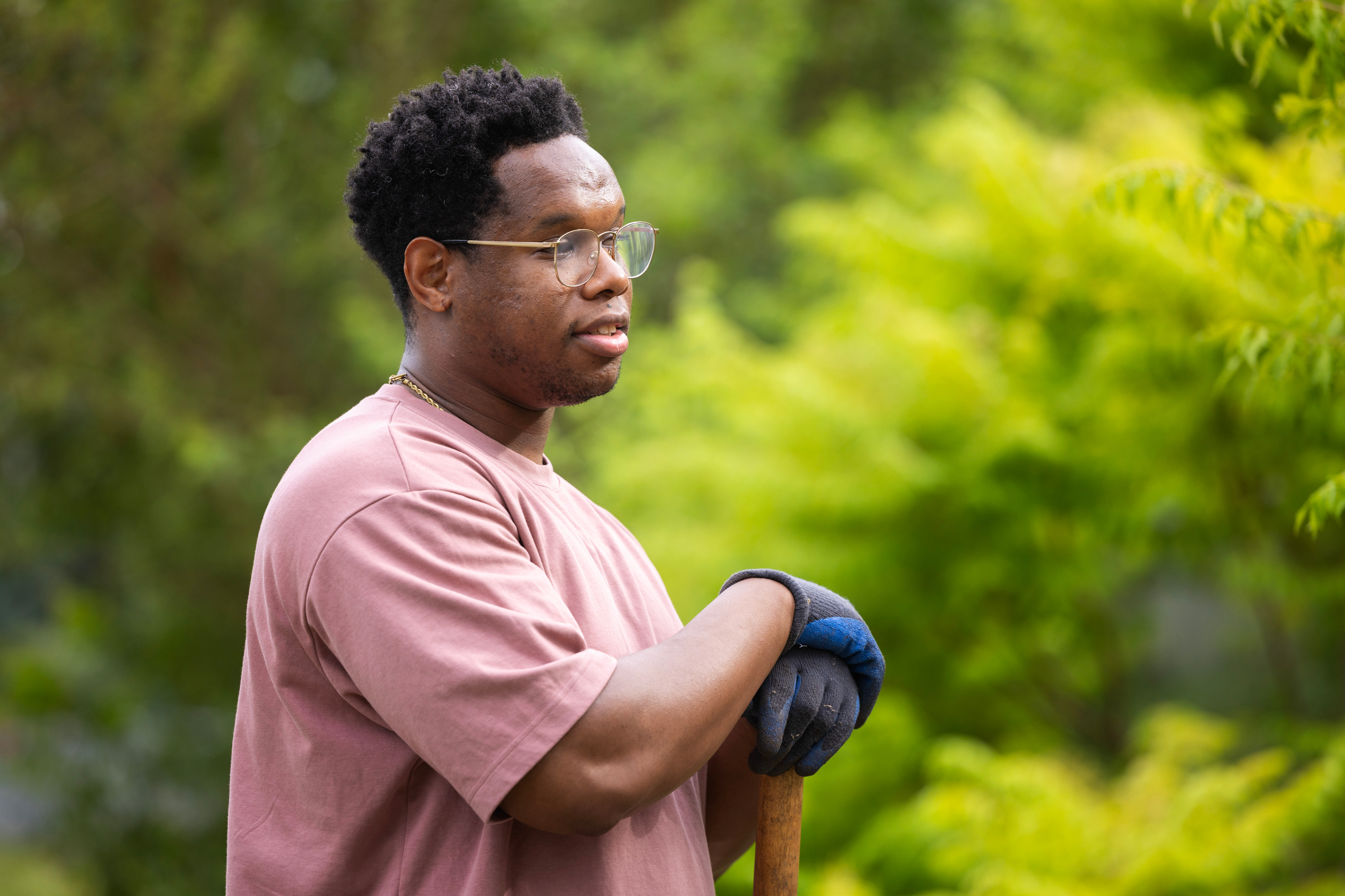 A man with black hair and gold-rimmed glasses wears a muted pink T-shirt and gardening gloves. His arms are crossed, and he leans on a wooden pole as he looks off frame toward the right. Behind him are green plants out of focus.