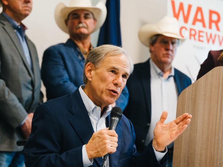 Texas Gov. Greg Abbott speaks into a microphone with his left hand raised in a gesture, addressing an audience. He is dressed in a navy suit jacket over a light-blue collared shirt.