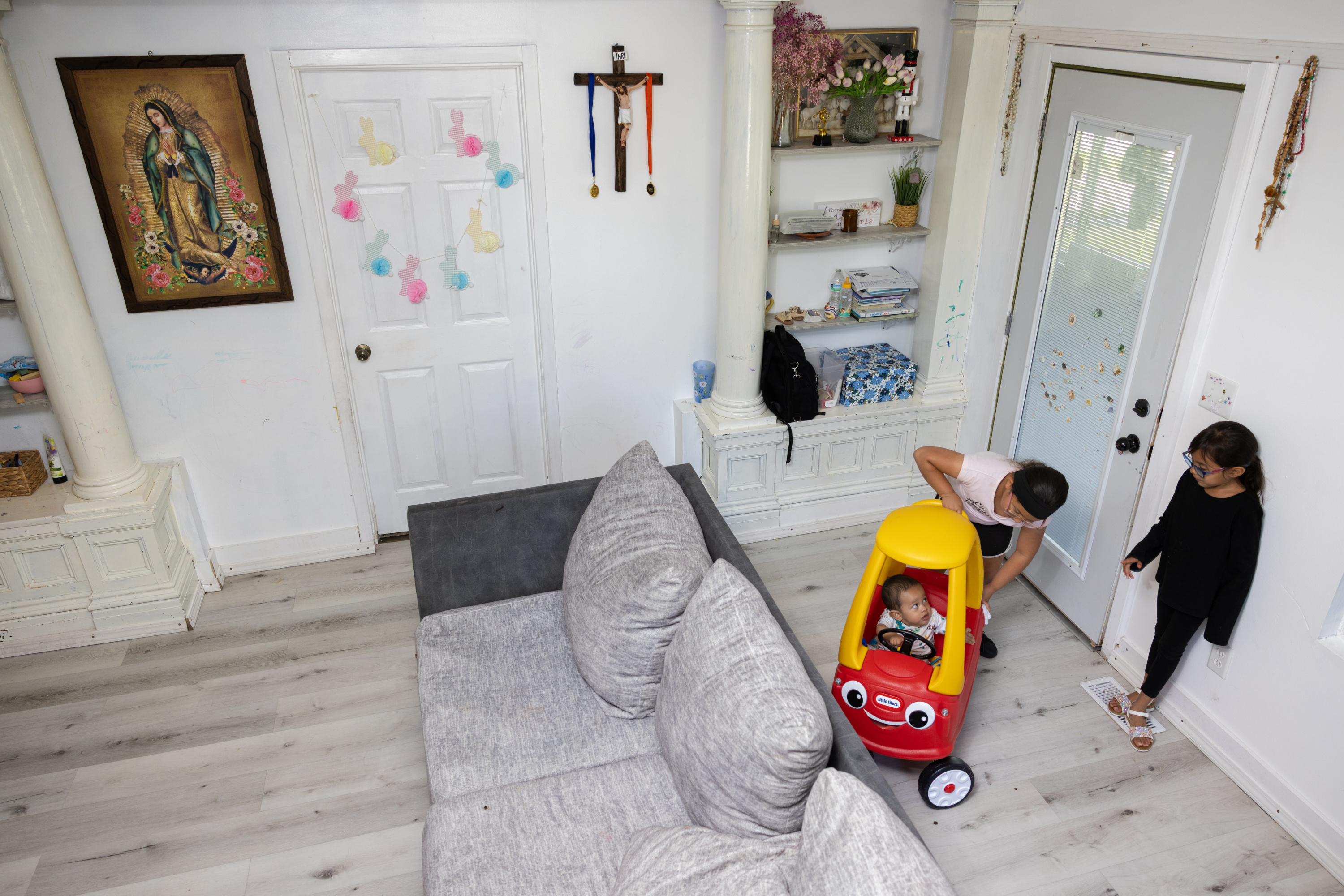Two small children look at a baby in a yellow and red toy car inside a gray, tidy living room. There is a crucifix on the wall with red and blue medallions hanging from it. A painting of the Virgin of Guadalupe also decorates the wall.