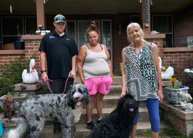 A man, two women and two dogs stand on the front steps in front of a brick house.