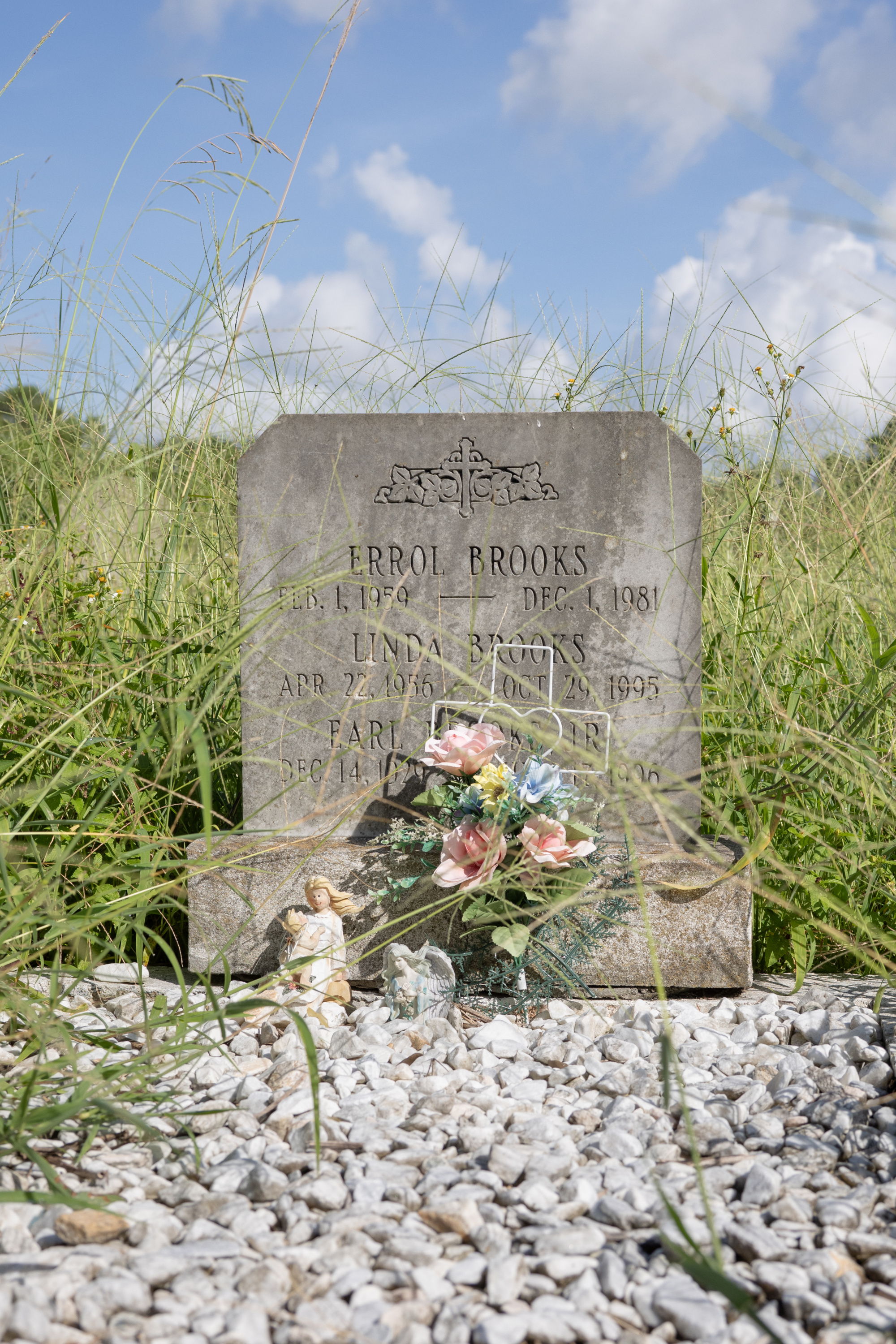 A gravestone with a cross etched on top and the words “Errol Brooks, Feb. 1 1959 — Dec. 1, 1981.” “Linda Brooks, Apr. 22, 1956 — Oct. 29, 1995,” and “Earl.” A bouquet of flowers and a cross obscure the last name. Two small angel statues also lean against the grave. Gravel sits in front of the grave, and long green grass grows behind the grave. The sky is blue with some clouds.