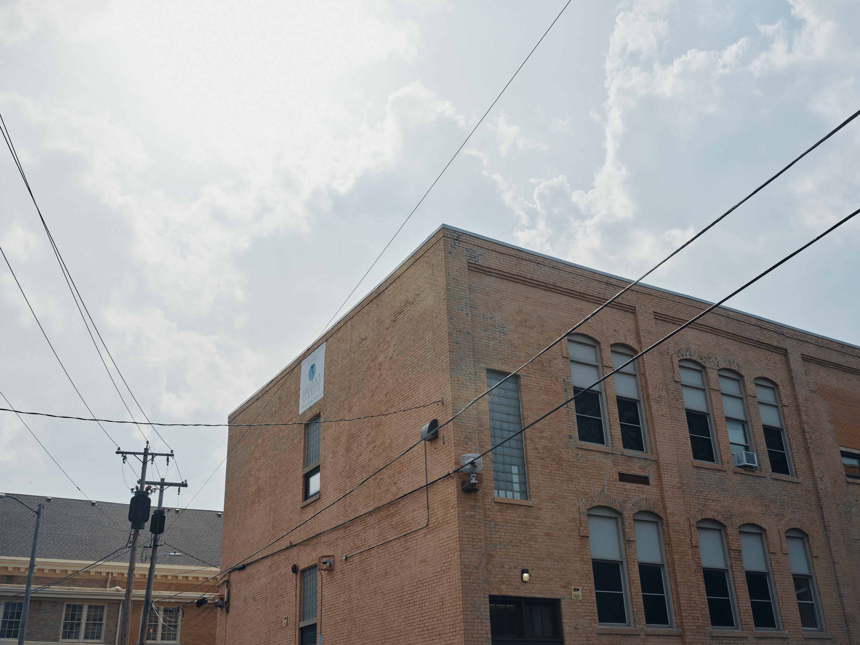 Power lines cross near a two-story, brown brick building with tall windows. The sky has large gray clouds.
