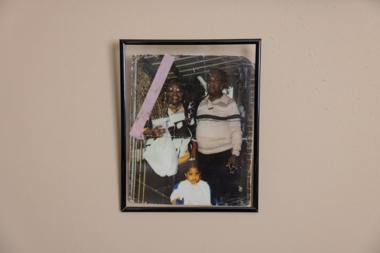 A framed photograph on a beige wall. The photograph has crease lines and a piece of purple tape on the top left corner. The photo shows a small child standing between two adults.