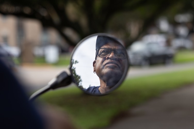 A circular mirror shows a man’s reflection. The man wears glasses, a baseball cap and an earring. The background is out of focus: a tree, a green lawn and cars parked outside of a building.
