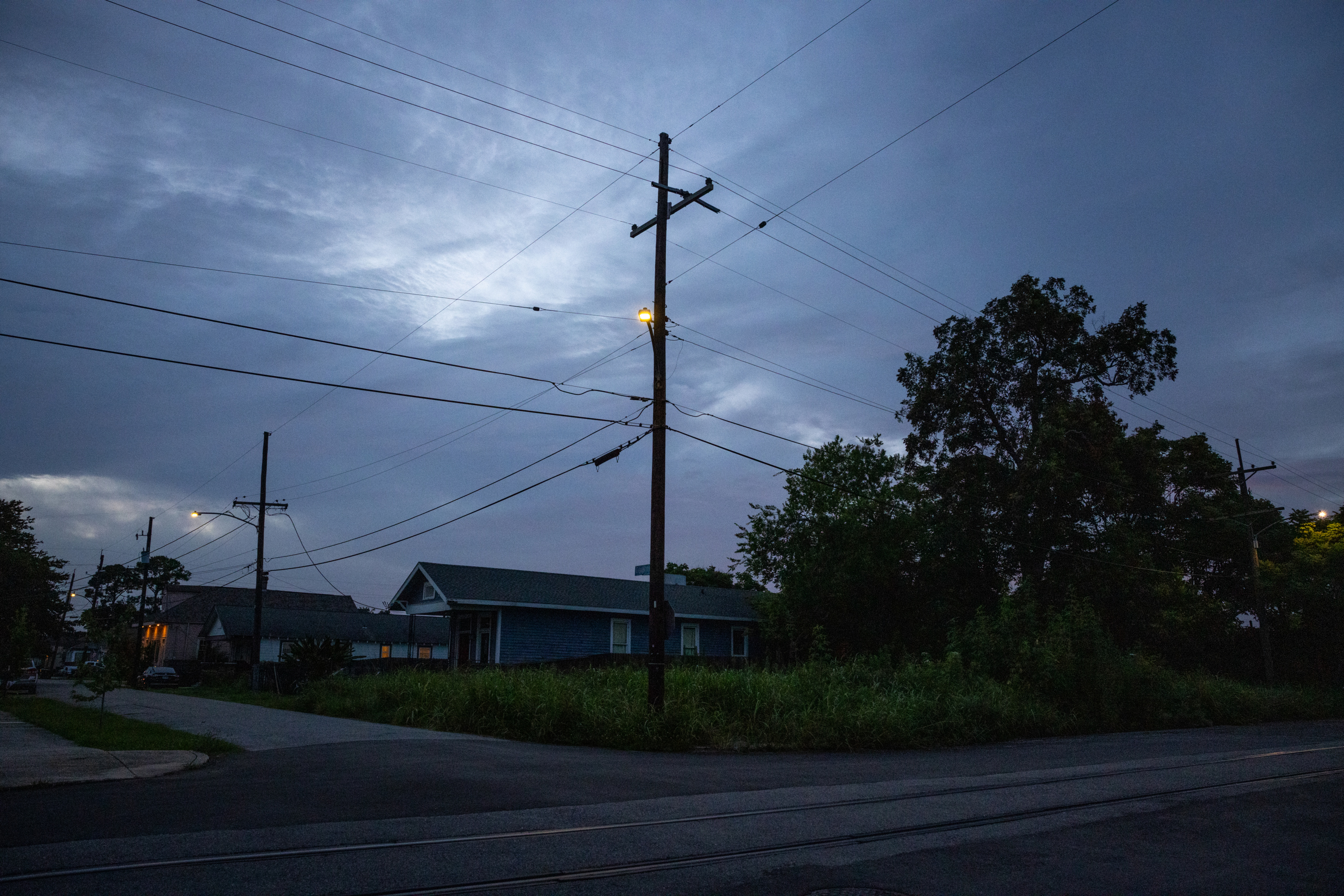 A dark, cloudy sky hangs over an intersection with silhouetted trees, power lines and one-story houses with some lit windows.