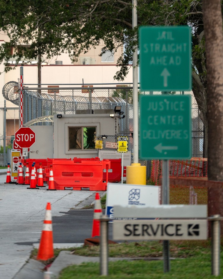 A secured jail entrance featuring a guard booth, red traffic barricades, orange traffic cones and green directional road signs.