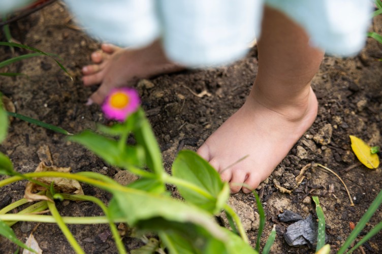 A toddler’s bare feet rest in soil. The photo is framed by flowers and leaves that are out of focus.