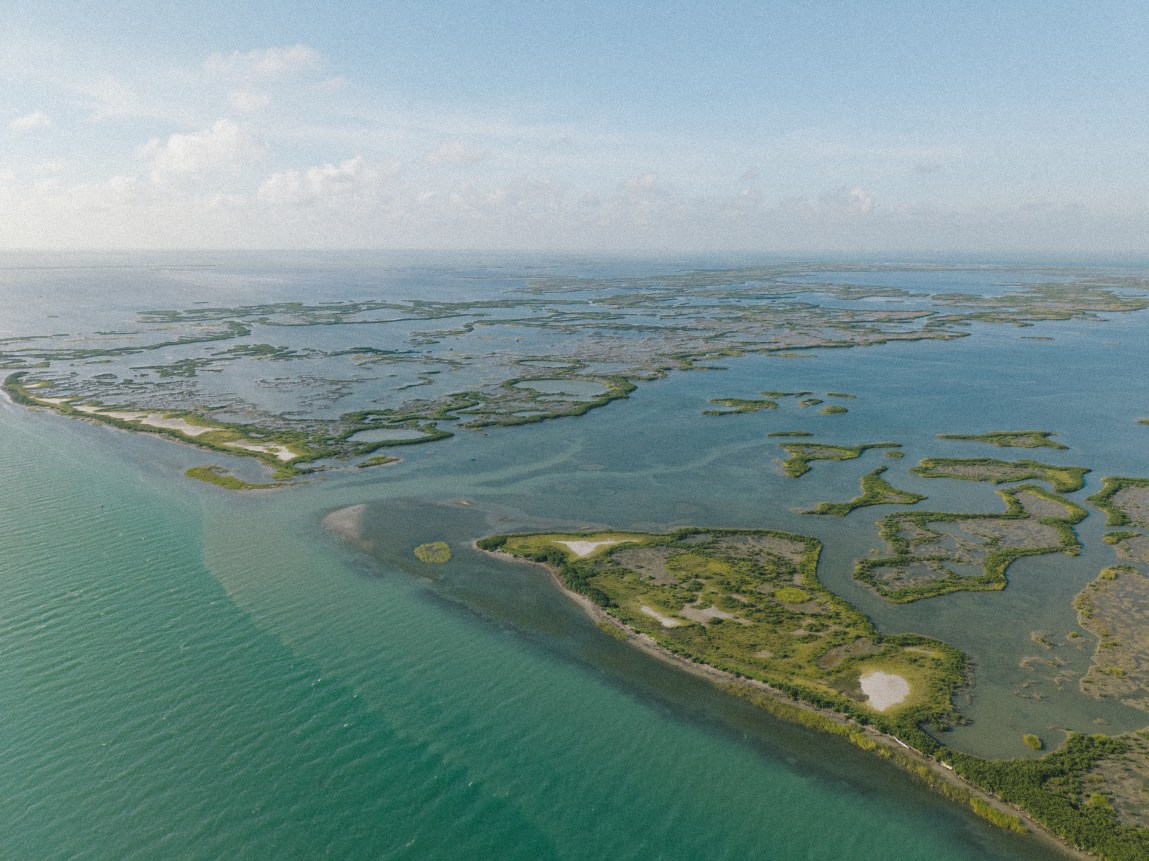 An aerial photograph captures a vast coastal marshland surrounded by vibrant turquoise and deep blue waters. Small green islands, winding estuaries and shallow mudflats stretch across the landscape.