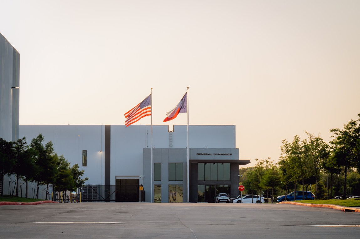 An American flag and a Texas flag displayed outside a gray and white building with a General Dynamics sign.