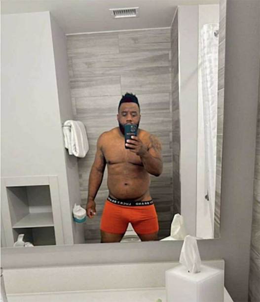 A man stands in a bathroom facing the mirror, holding up his phone. He is wearing only boxer briefs, pulled down slightly at the waist on one side.
