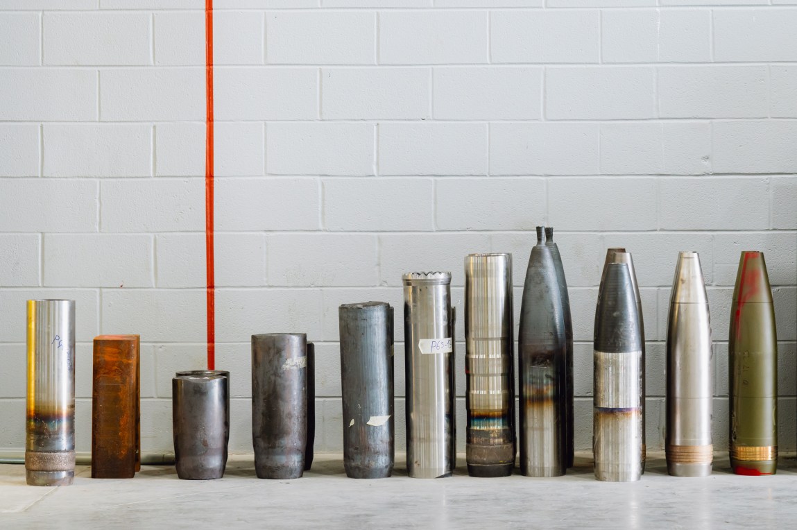 A row of artillery shells at various stages of production in front of a cement wall painted white.