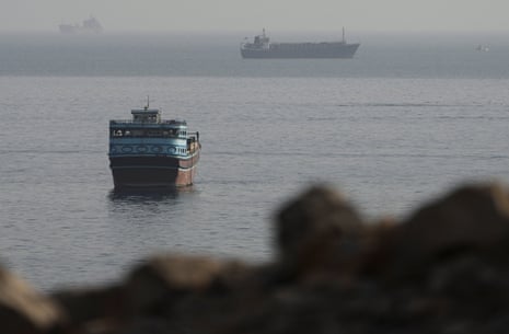 Image shows a small ship in the foreground with larger vessels in the background in the strait of Hormuz. 