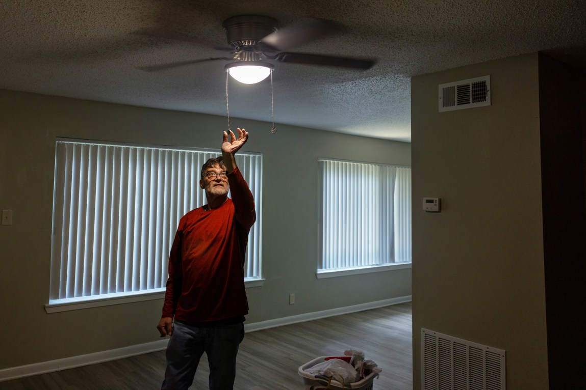 A man reaches up and pulls the light switch to an overhead light in a simple apartment room.