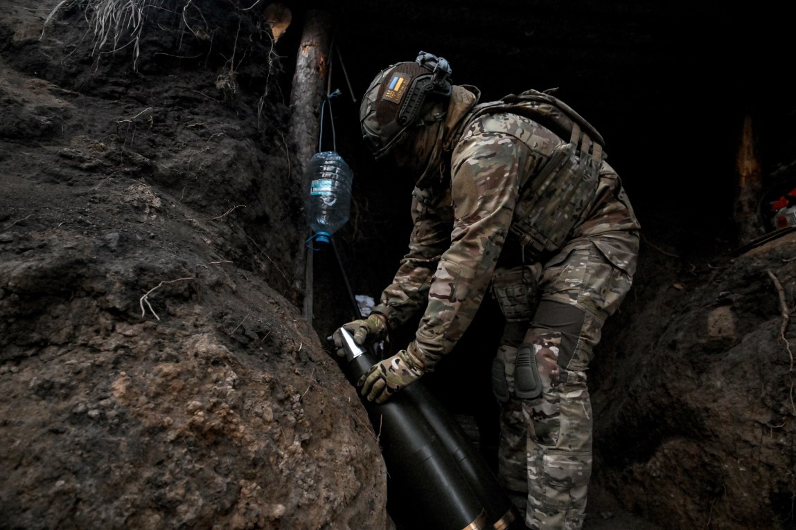 A soldier in a camouflage uniform handles 155 mm artillery shells, which are black with shiny, pointed tips.