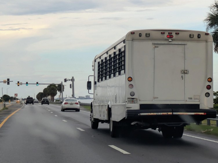 A white transport van with barred windows driving on a multi-lane highway near the coast.