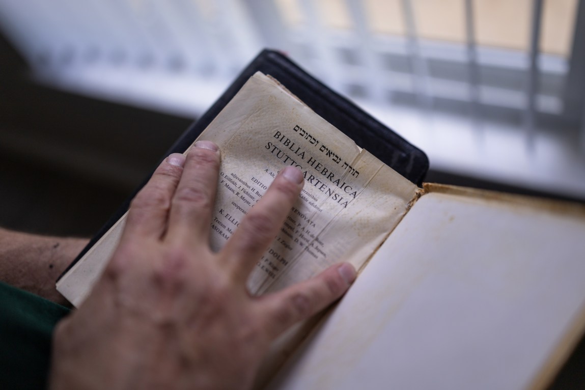 A man’s hand holds a book open to a page that reads “Biblica Hebraica.”