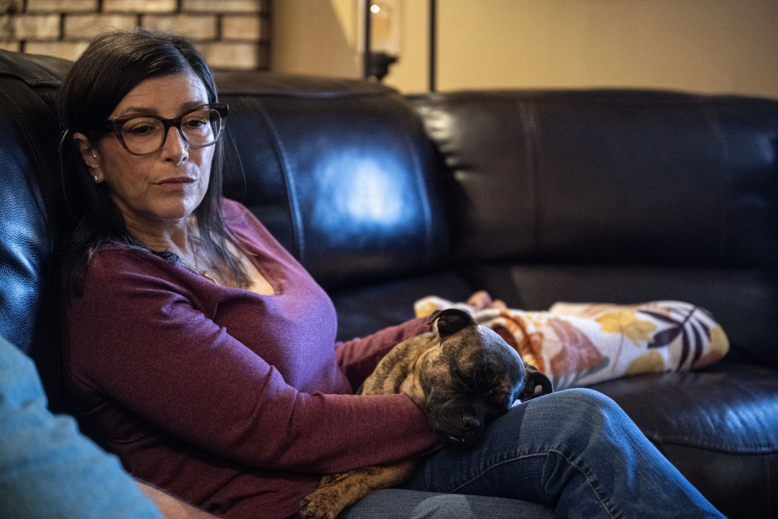 A woman wearing a long-sleeved purple shirt and dark-rimmed glasses holds a small dog in her lap. She sits on a leather couch in a room with yellow walls.