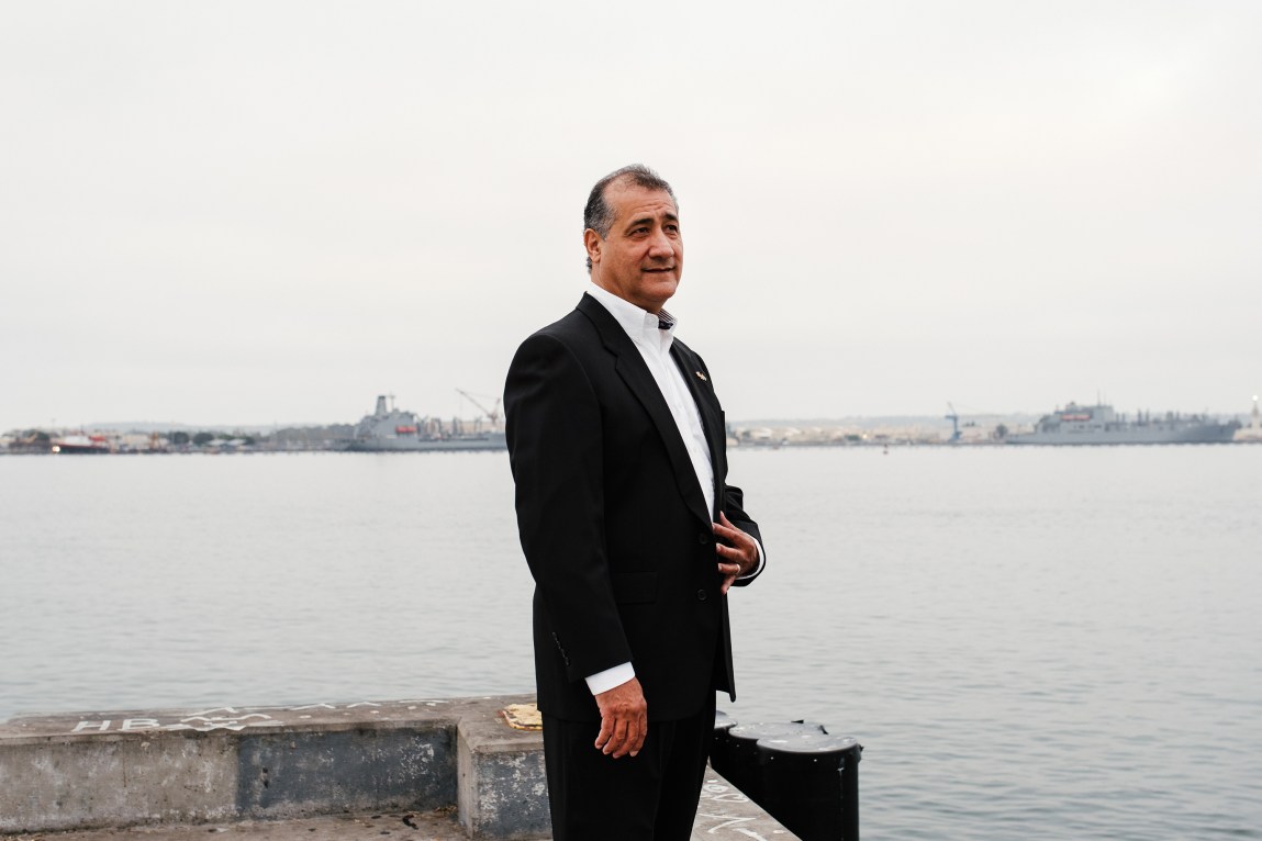 A man in business attire stands on a pier next to a large body of water. Ships can be seen in the distance.