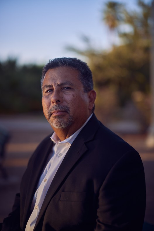 A middle-aged man in a dark suit jacket and white shirt looks directly toward the viewer in a portrait made at dusk in a community park.