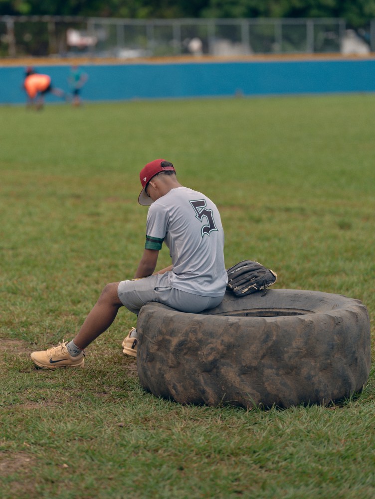 A young baseball player wearing No. 5 sits with his head down on a large tire at the edge of a baseball field.