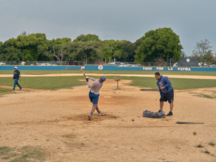A baseball player completes a swing during batting practice on a dirt field with a coach watching nearby.