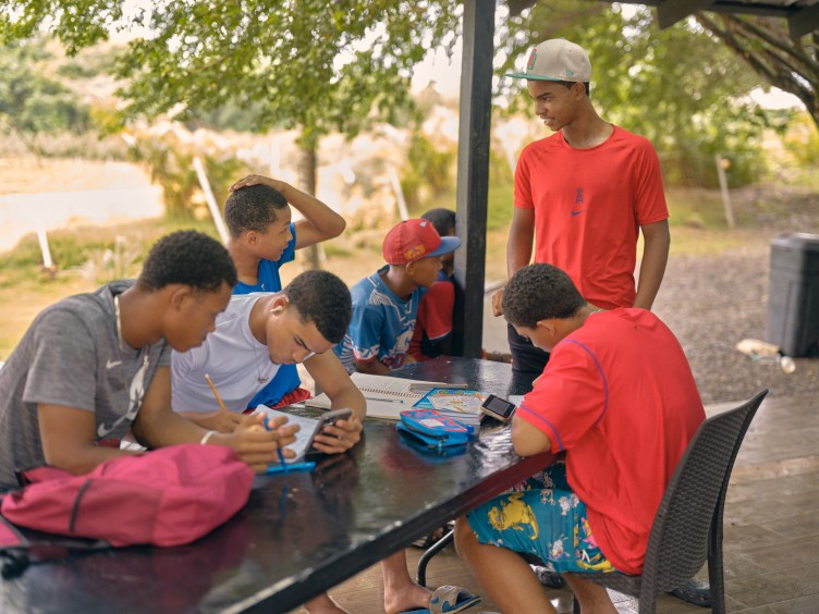 A group of young male baseball players sit around a dark picnic table under a covered outdoor shelter studying, writing in notebooks and looking at phones.