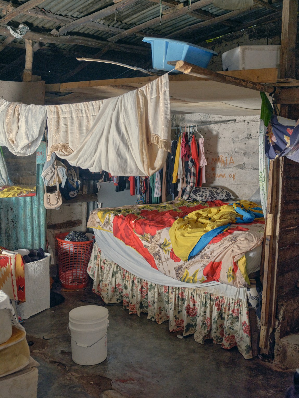 The interior of a bedroom with a brightly blanketed bed, hanging laundry, and a corrugated metal and wood ceiling structure.