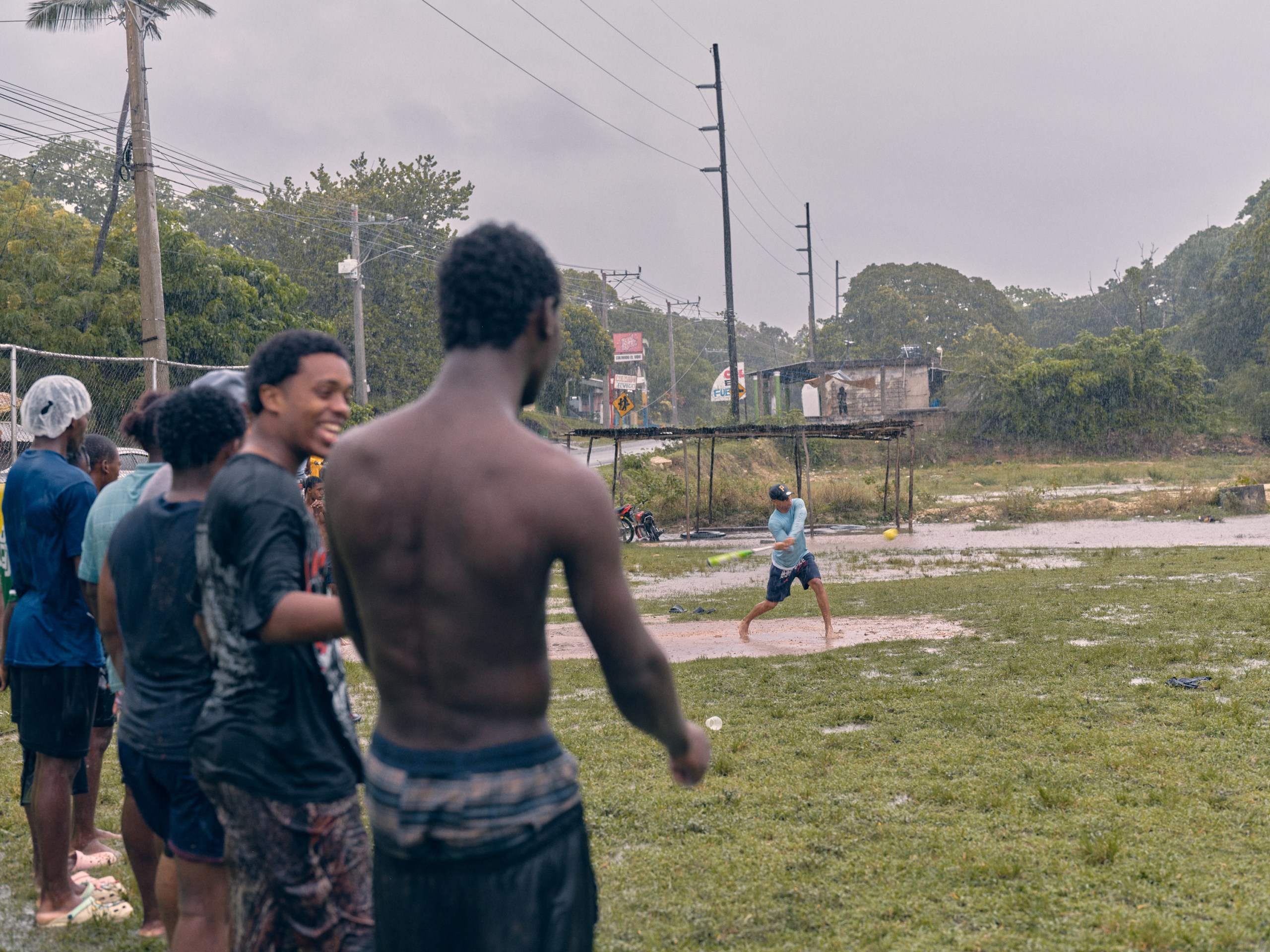 Young men gather outdoors in the rain to watch a barefoot batter swing at a yellow ball on a flooded, muddy field.