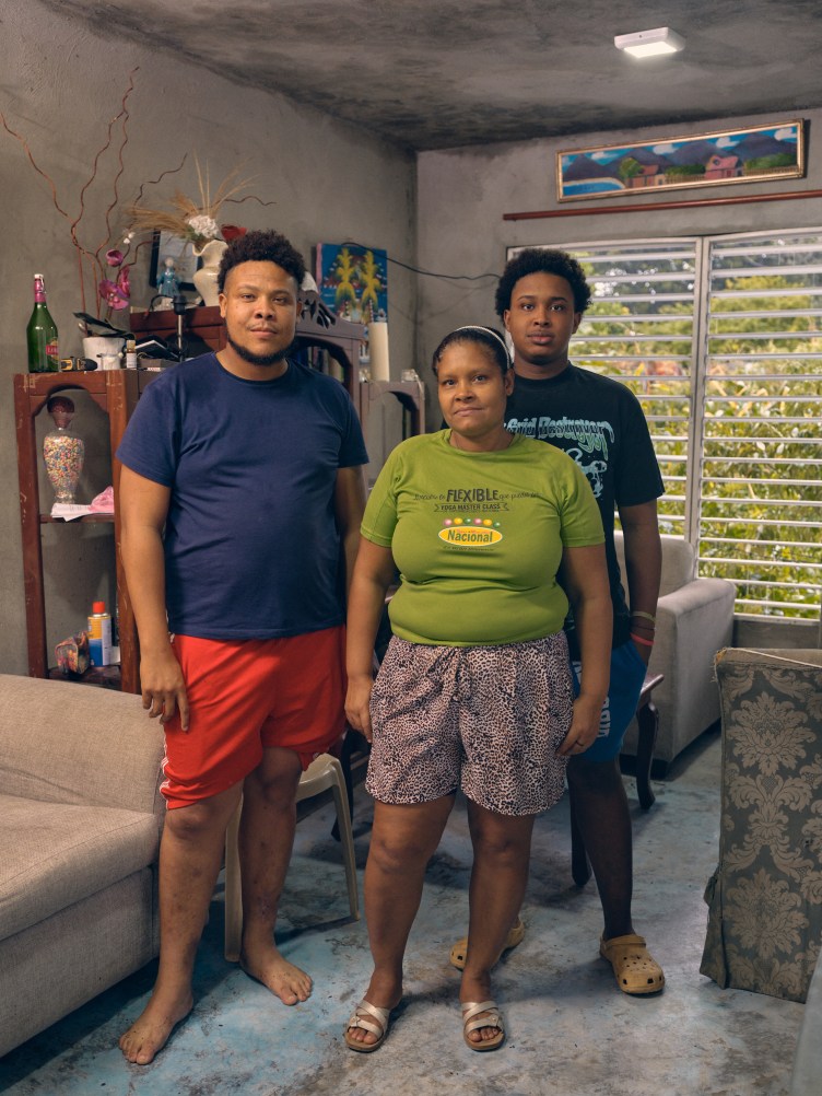 Two young men stand behind a woman, all looking at the camera. Behind them is a bright living room with colorful flowers and paintings.