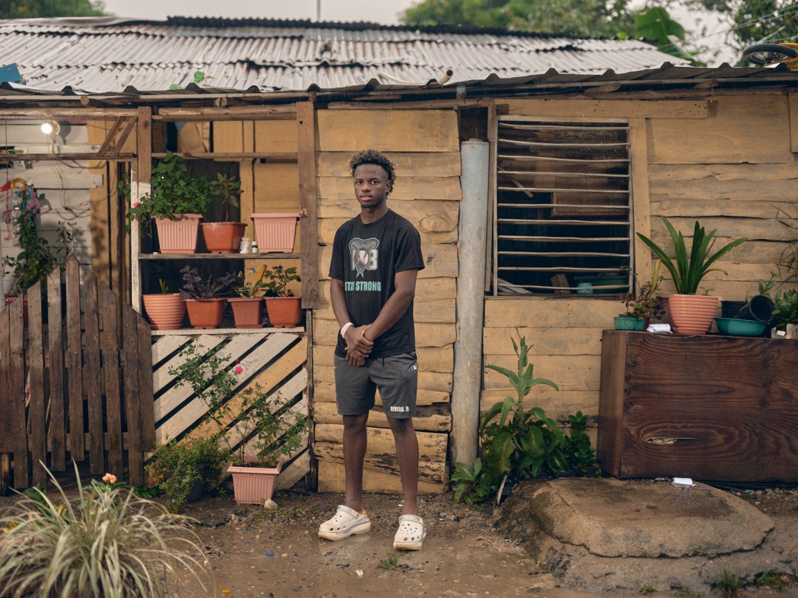 A young man wearing a black T-shirt and white clogs stands outside a rustic wooden house surrounded by potted plants.