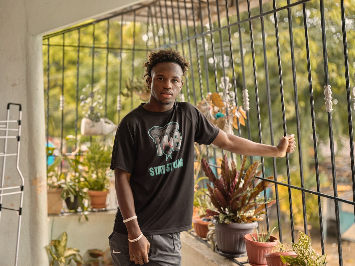 A young man wearing a black “stay strong” T-shirt stands on a porch next to potted plants, holding onto a decorative metal security gate.