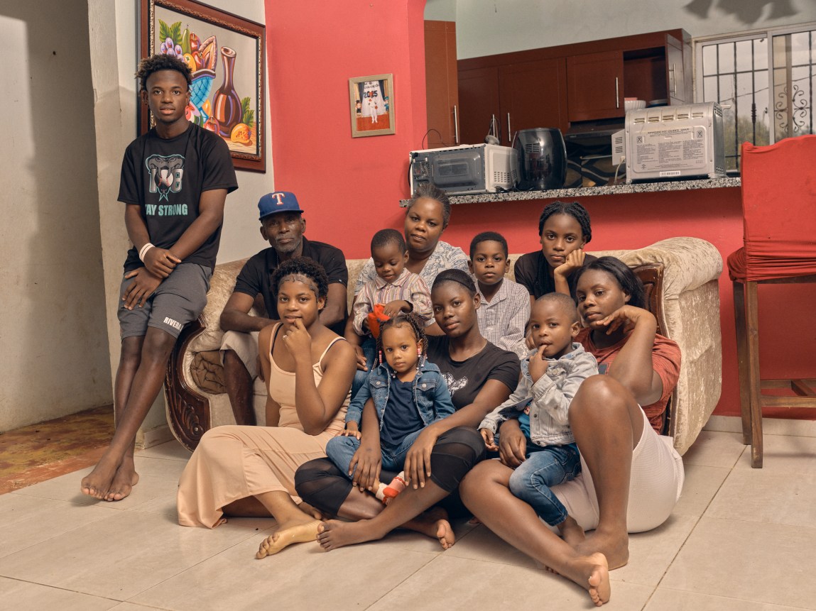 A large, multigenerational family poses together for a portrait in their bright living room, seated on a couch and the floor.