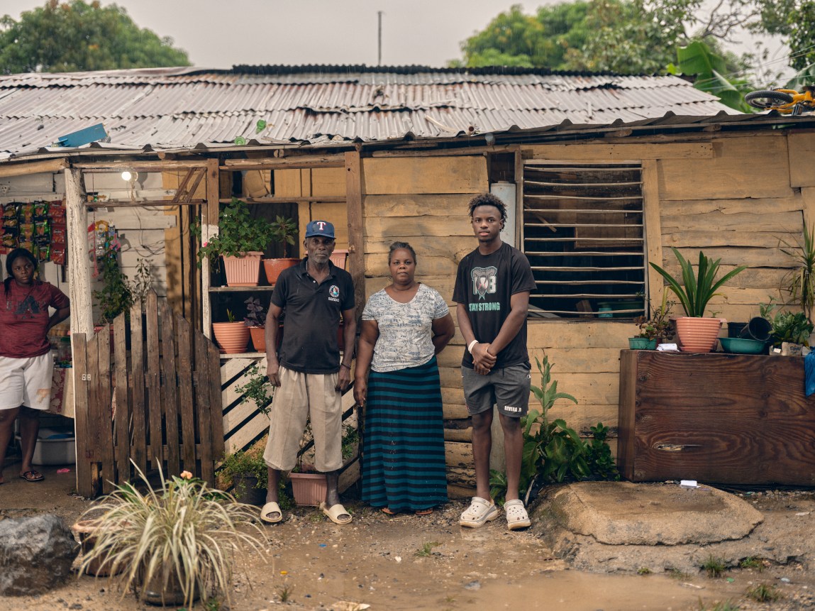 A family of three stands outside their wooden, corrugated metal-roofed home decorated with potted plants.