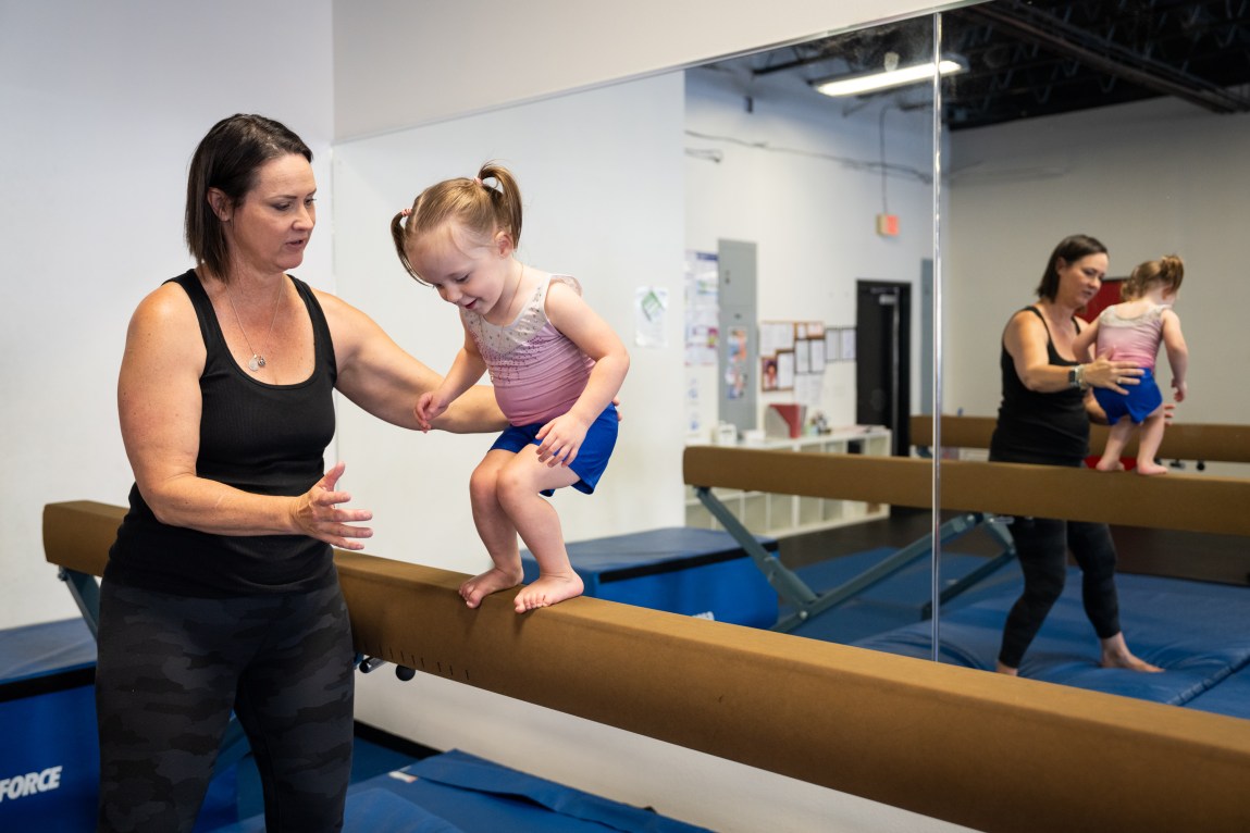 A woman spotting a young girl walking on a low balance beam in a gymnastics studio, with their reflection visible in a large mirror behind them.