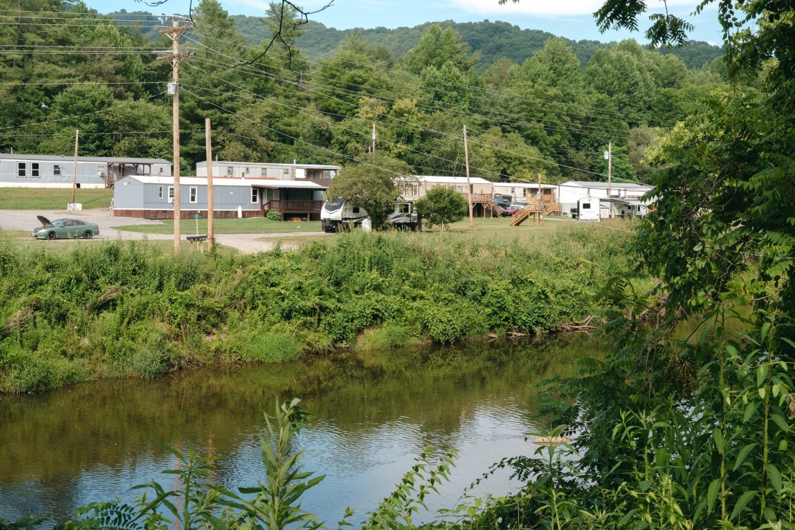 Single-wide trailer homes, campers and cars can be seen on the other side of a river. Lush green vegetation grows around the river and park site.