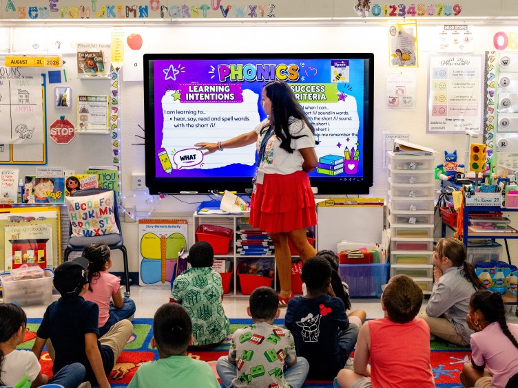 A woman points to a screen displaying a phonics lesson at the front of an elementary school classroom. Boys and girls sit cross-legged on the floor in front of her.