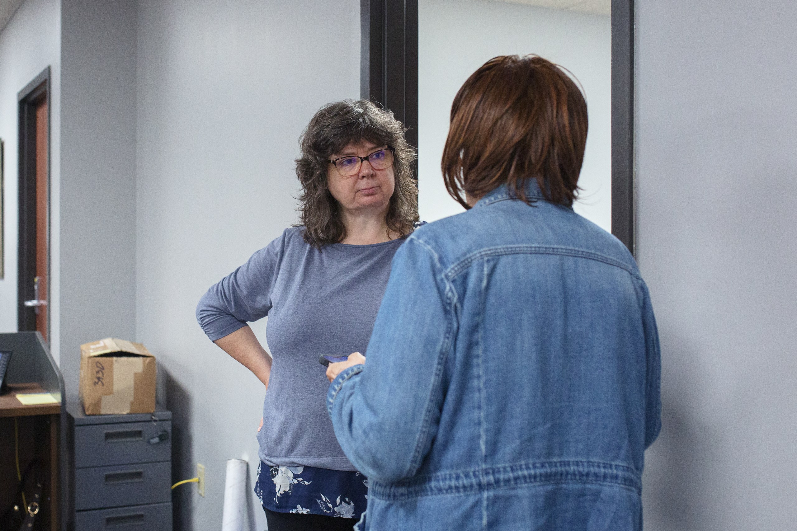 A middle-aged woman wearing glasses and a long-sleeved gray shirt faces the viewer while speaking with another woman whose back is to the camera. Both are standing in a doorway, inside a nondescript light gray office space.