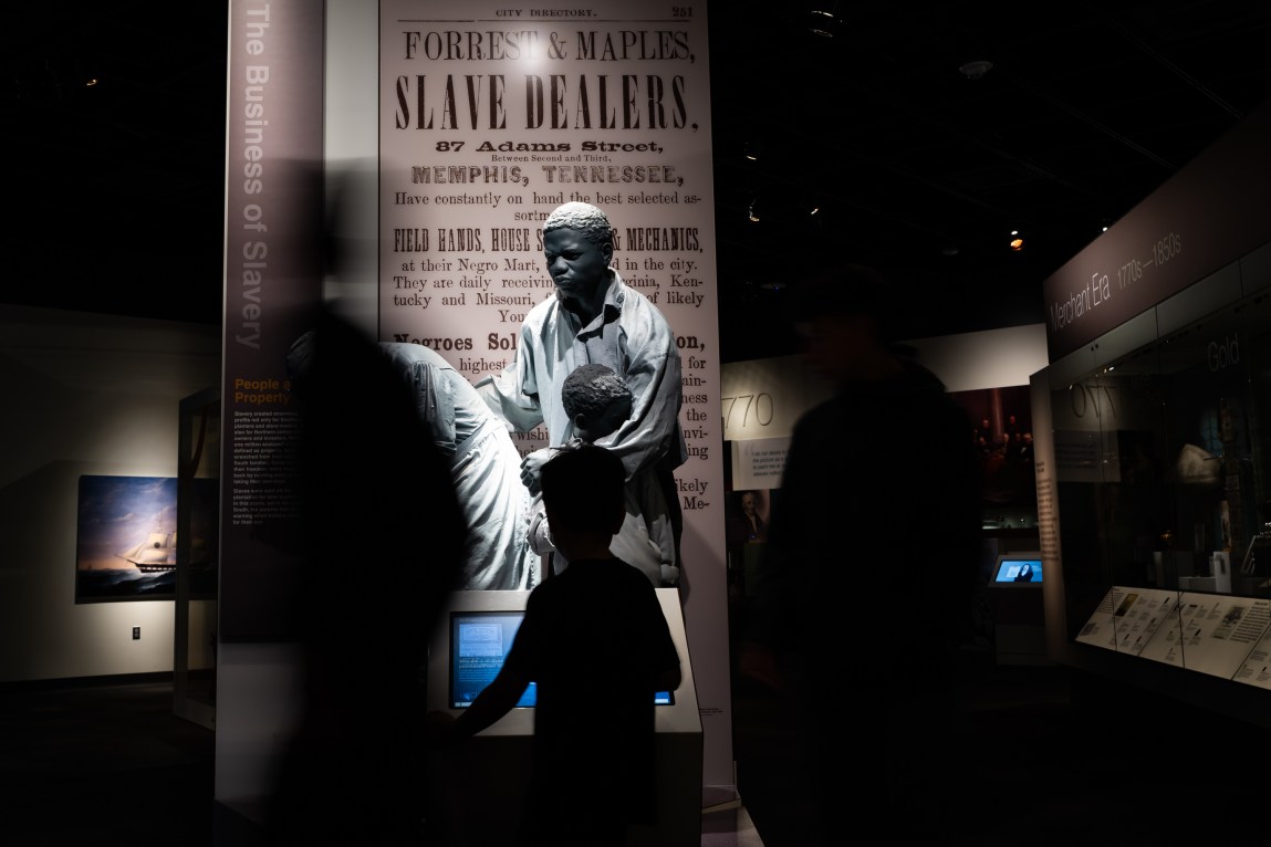 People in front of a museum exhibit showing an advertisement that says “Forrest & Maples, slave dealers,” with a Memphis, Tennessee, address.
