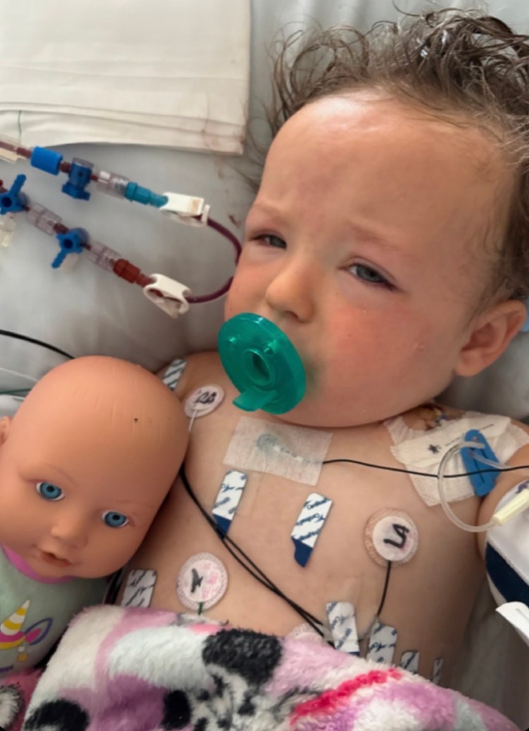A toddler in a hospital bed with a green pacifier and medical electrodes attached to her chest, with a baby doll resting next to her.