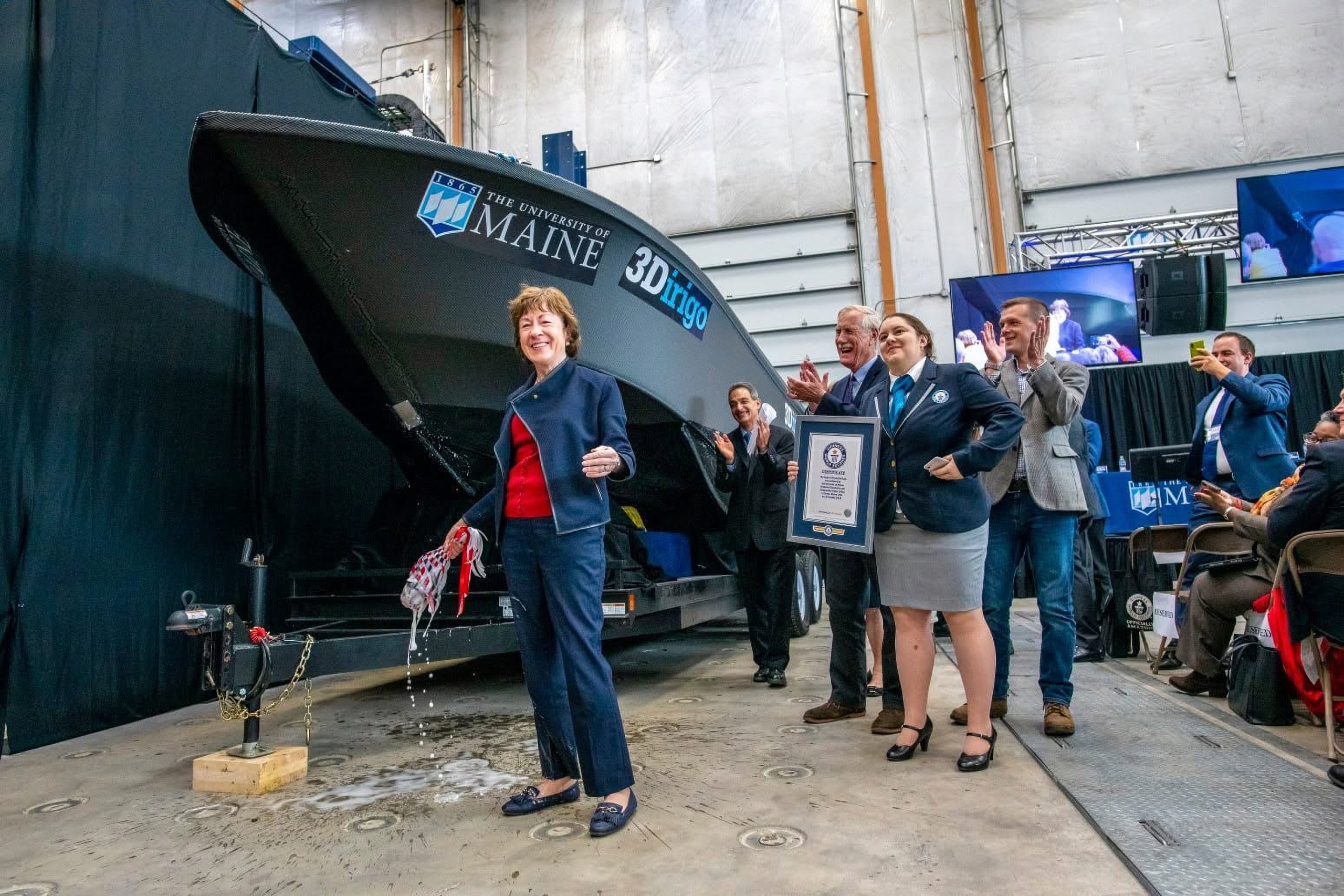 A woman in a blue suit and red shirt stands in front of a boat while holding a broken champagne bottle during a publicity event.