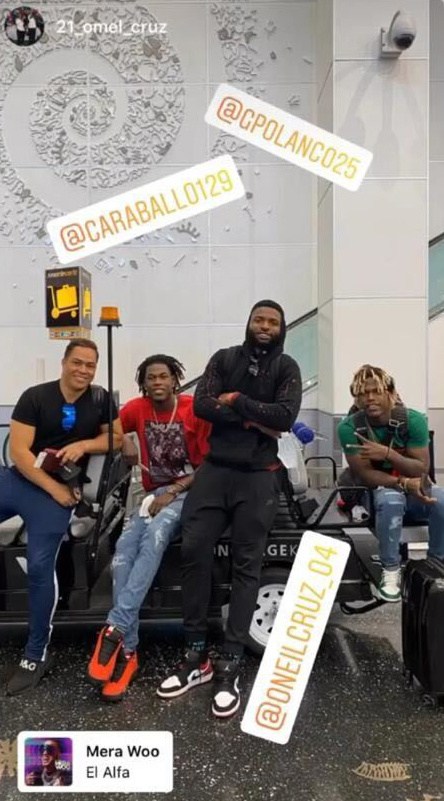 Four men pose together around an airport utility cart, featuring social media handle overlays and a music tag for “Mera Woo” by El Alfa.