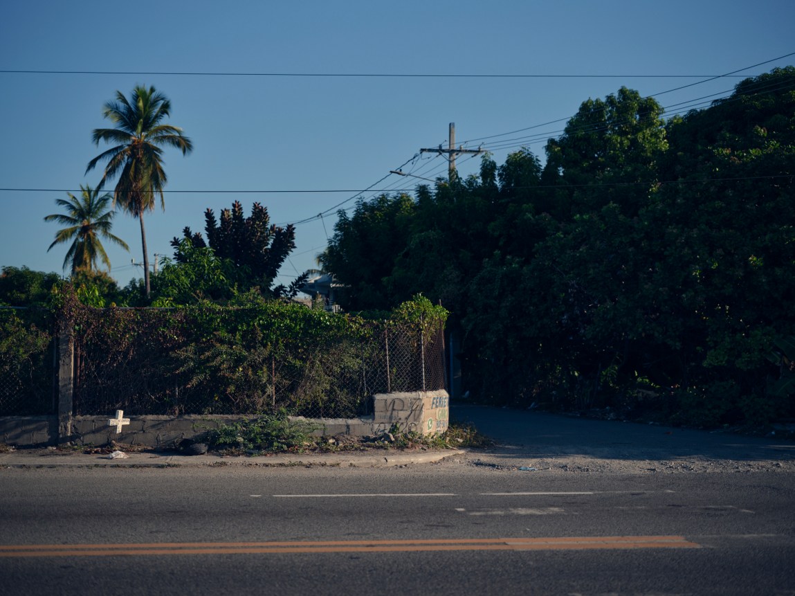 An empty road with lush green trees and power lines behind it. On the sidewalk is a white cross.
