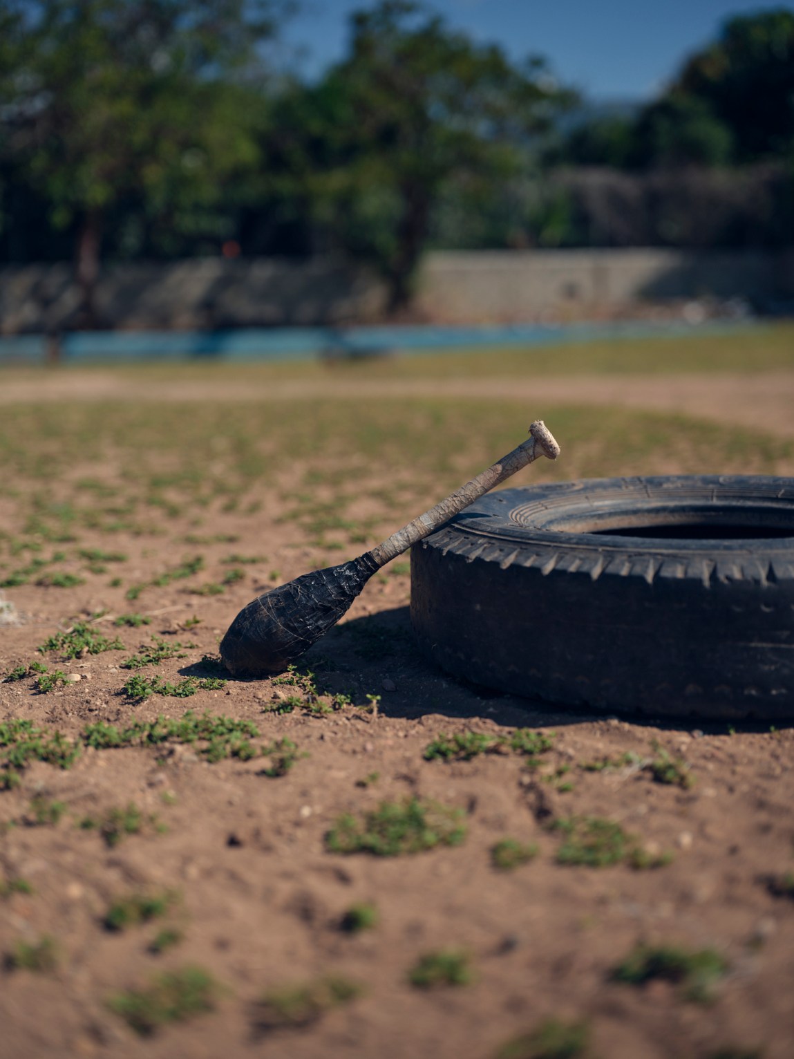 A worn baseball bat wrapped in dark tape resting against a large rubber tire on a dirt field.