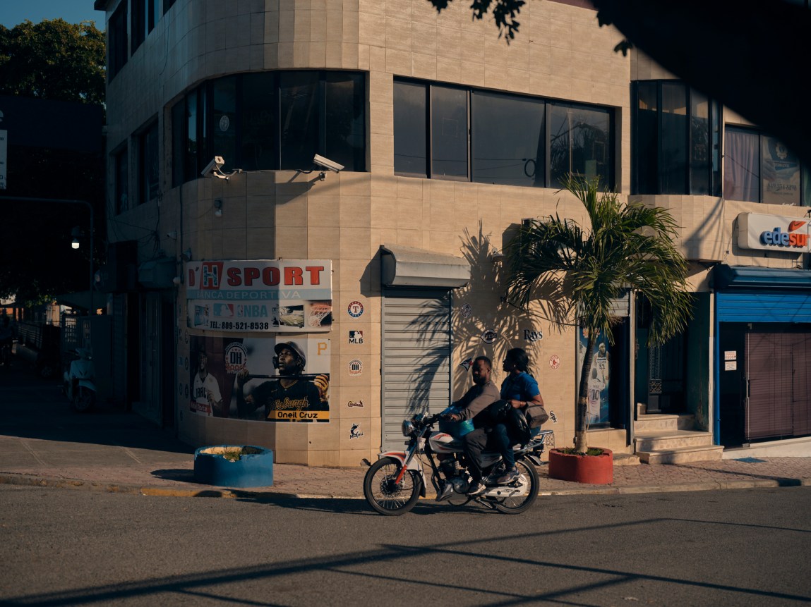 Two people sit on a motorcycle on a street bathed in warm light. Behind them there is a building with posters, one with a photograph of Oneil Cruz. On the sidewalk sits a palm tree.
