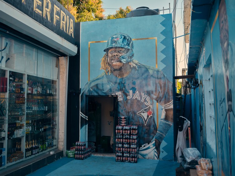 On a blue wall is a mural of Vladimir Guerrero Jr. in a Blue Jays jersey and helmet. He has his tongue sticking out to the side in concentration. In front of the mural are a doorway and a large stack of bottles wrapped in plastic.