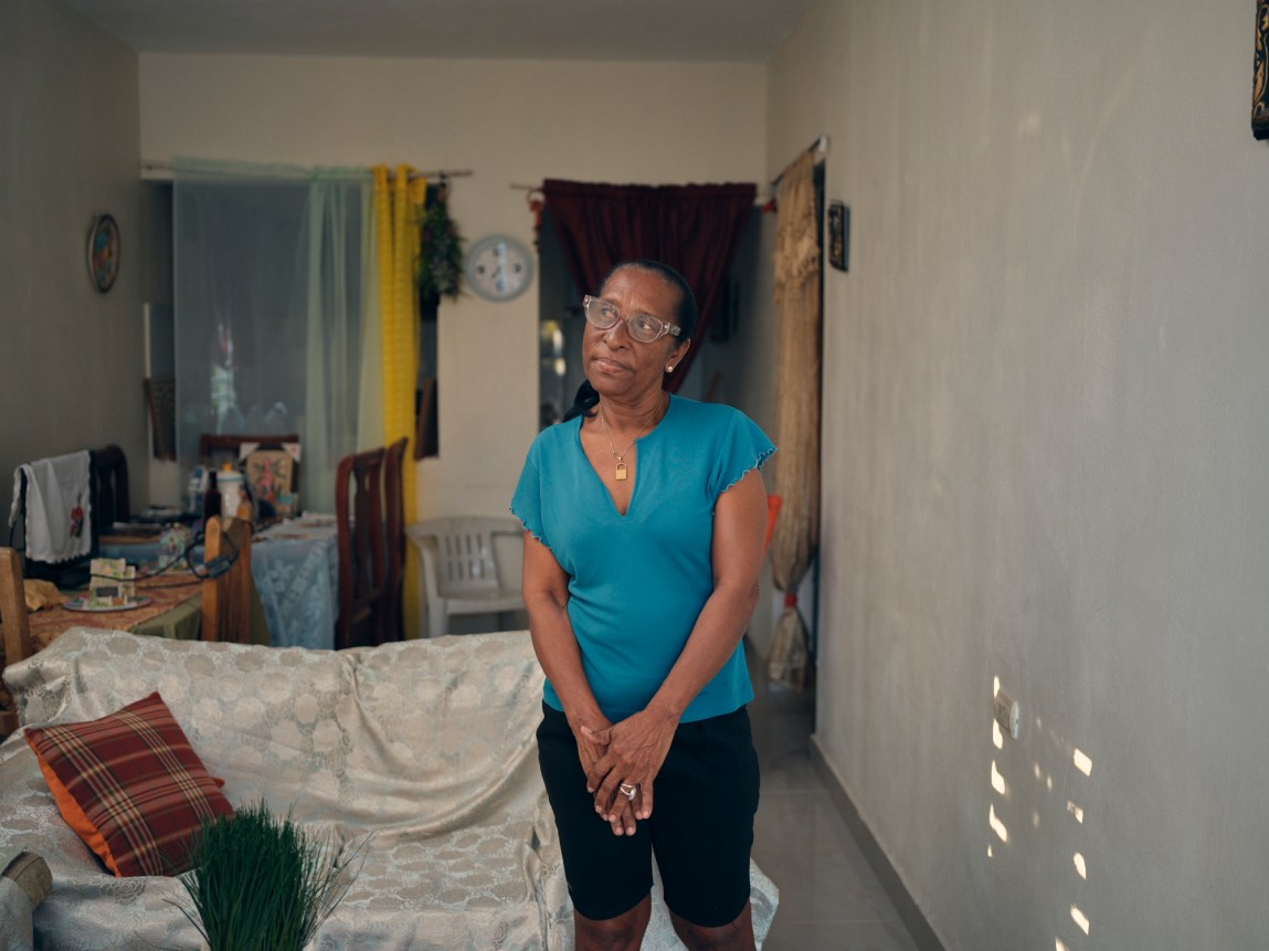A woman in a bright blue shirt wearing glasses looks to her right. Behind her is a couch with a white patterned cloth draped on it and a dinner table.