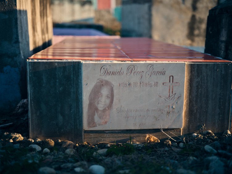 A grave with a photograph of a young woman smiling on it. Beside her it reads “Daniela Perez Garcia” with an illustration of a cross and a dove.