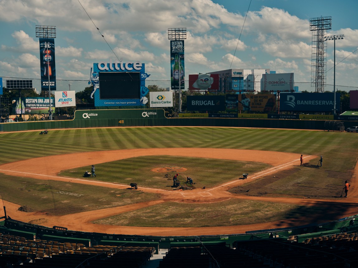 A high-angle view of a professional baseball stadium as groundskeepers maintain the infield dirt and grass under a partly cloudy sky.